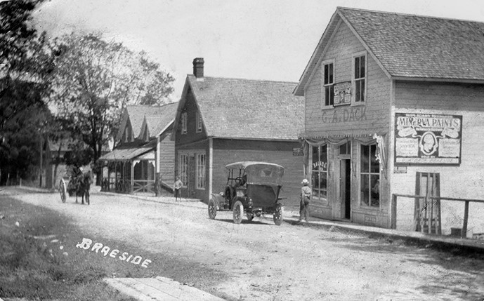 Carte postale d’une scène de rue de Braeside, avec quelques villageois, des commerces, une calèche et une voiture ancienne.