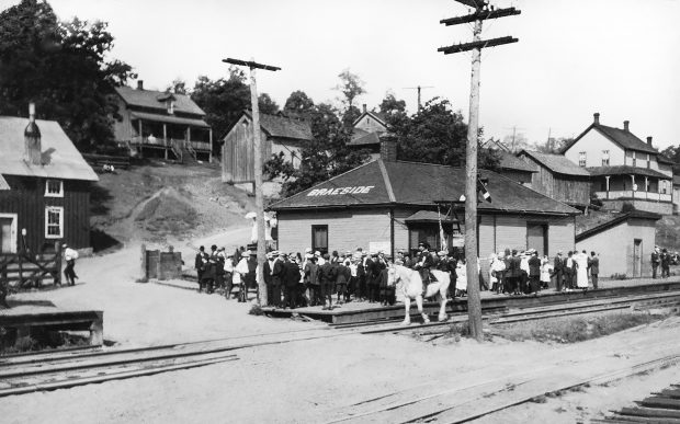 Une foule rassemblée devant la gare de train de Braeside regarde le défilé des orangistes mené par un homme sur un cheval blanc.
