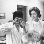 Photo en noir et blanc où deux femmes en sarrau de laboratoire examinent une fiole tenue par l’une d’entre elles.