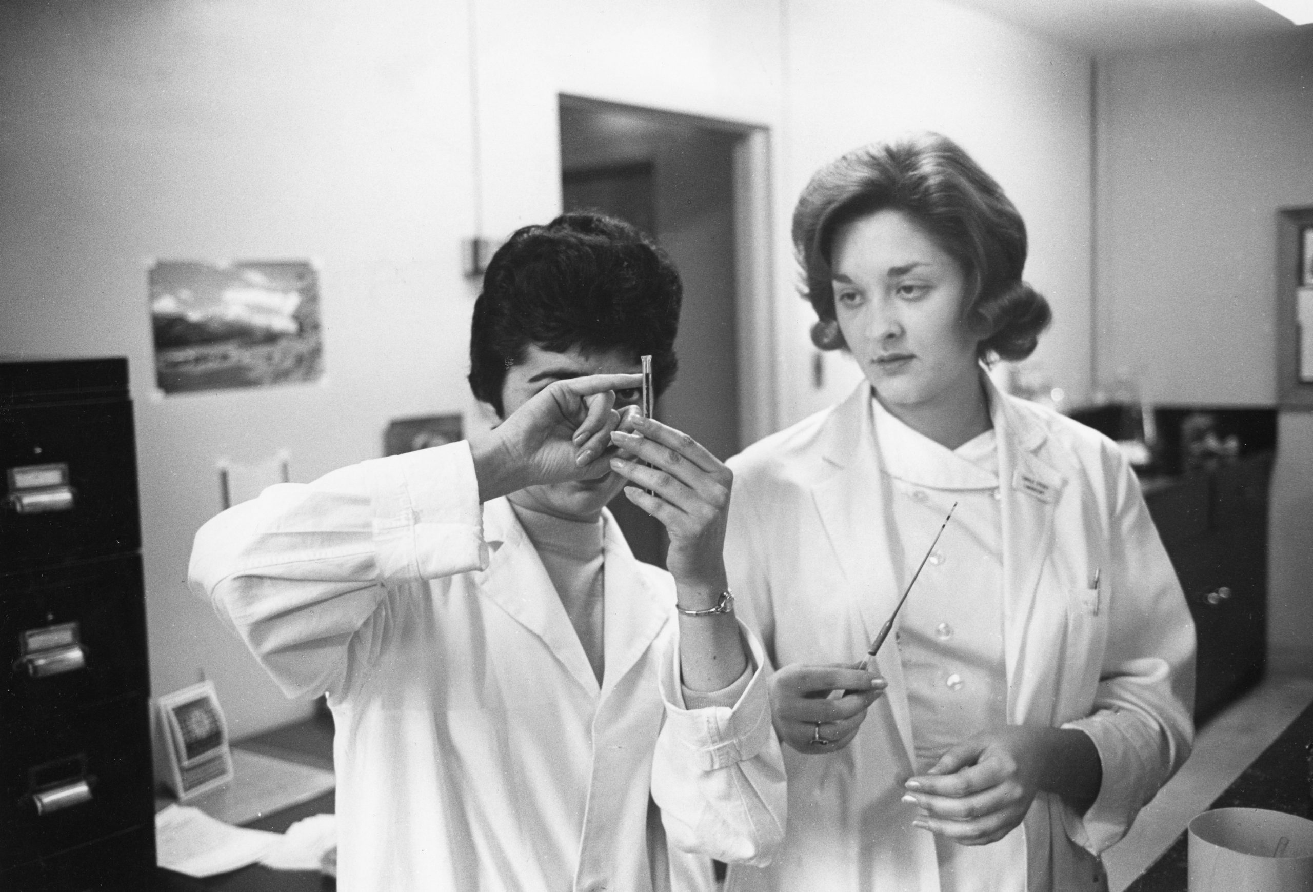 Photo en noir et blanc où deux femmes en sarrau de laboratoire examinent une fiole tenue par l’une d’entre elles.