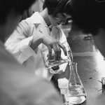 Photo en noir et blanc où une femme en sarrau de laboratoire transvase un liquide clair d’un bécher à un autre. Elle est encadrée par deux femmes plus âgées à l’avant-plan.
