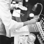 Photo en noir et blanc d’une femme en sarrau de laboratoire travaillant avec des fioles et des pipettes dans un laboratoire.