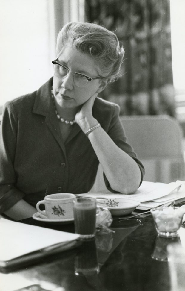 Photo en noir et blanc d’une femme en blazer assise à une table. Devant elle se trouvent des papiers et les restes de son thé. Elle supporte sa tête avec son bras pendant qu’elle écoute quelque chose que l’on ne voit pas.