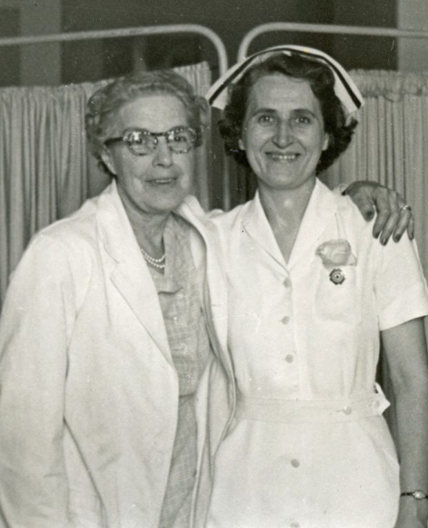 Photo en noir et blanc de deux femmes, dont une portant un sarrau de laboratoire. Celle-ci enlace l’autre femme en uniforme d’infirmière.