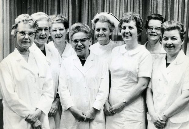 Photo en noir et blanc où huit femmes d’âge mûr en sarrau de laboratoire sourient en regardant l’appareil photo.