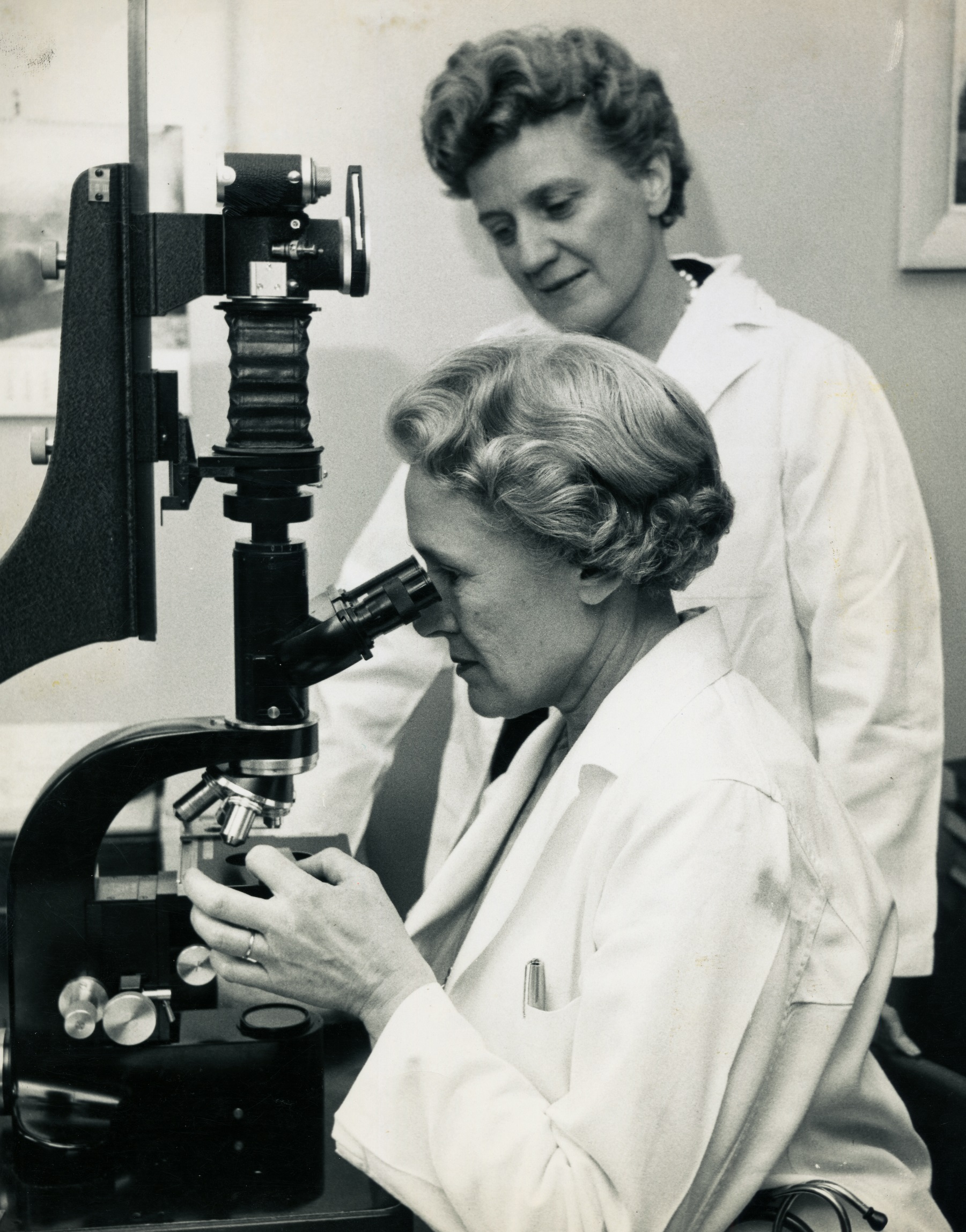 Photo en noir et blanc de deux femmes en sarrau de laboratoire. L’une d’entre elles regarde dans un grand microscope pendant que l’autre l’observe.