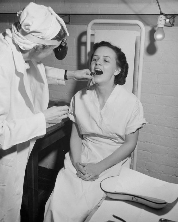 Photo en noir et blanc où une femme portant une blouse chirurgicale et un miroir frontal examine la bouche ouverte d’une jeune femme en chemise d’hôpital.
