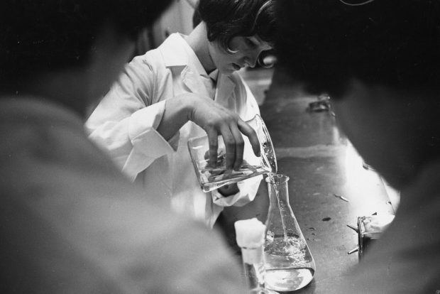 Photo en noir et blanc où une femme en sarrau de laboratoire transvase un liquide clair d’un bécher à un autre. Elle est encadrée par deux femmes plus âgées à l’avant-plan.
