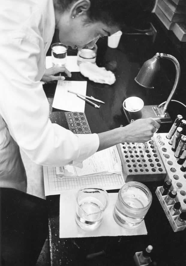Photo en noir et blanc d’une femme en sarrau de laboratoire travaillant avec des fioles et des pipettes dans un laboratoire.