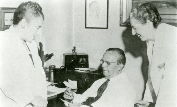 Photo en noir et blanc où deux femmes en sarrau de laboratoire parlent à un homme d’âge mûr portant une chemise élégante et une cravate.