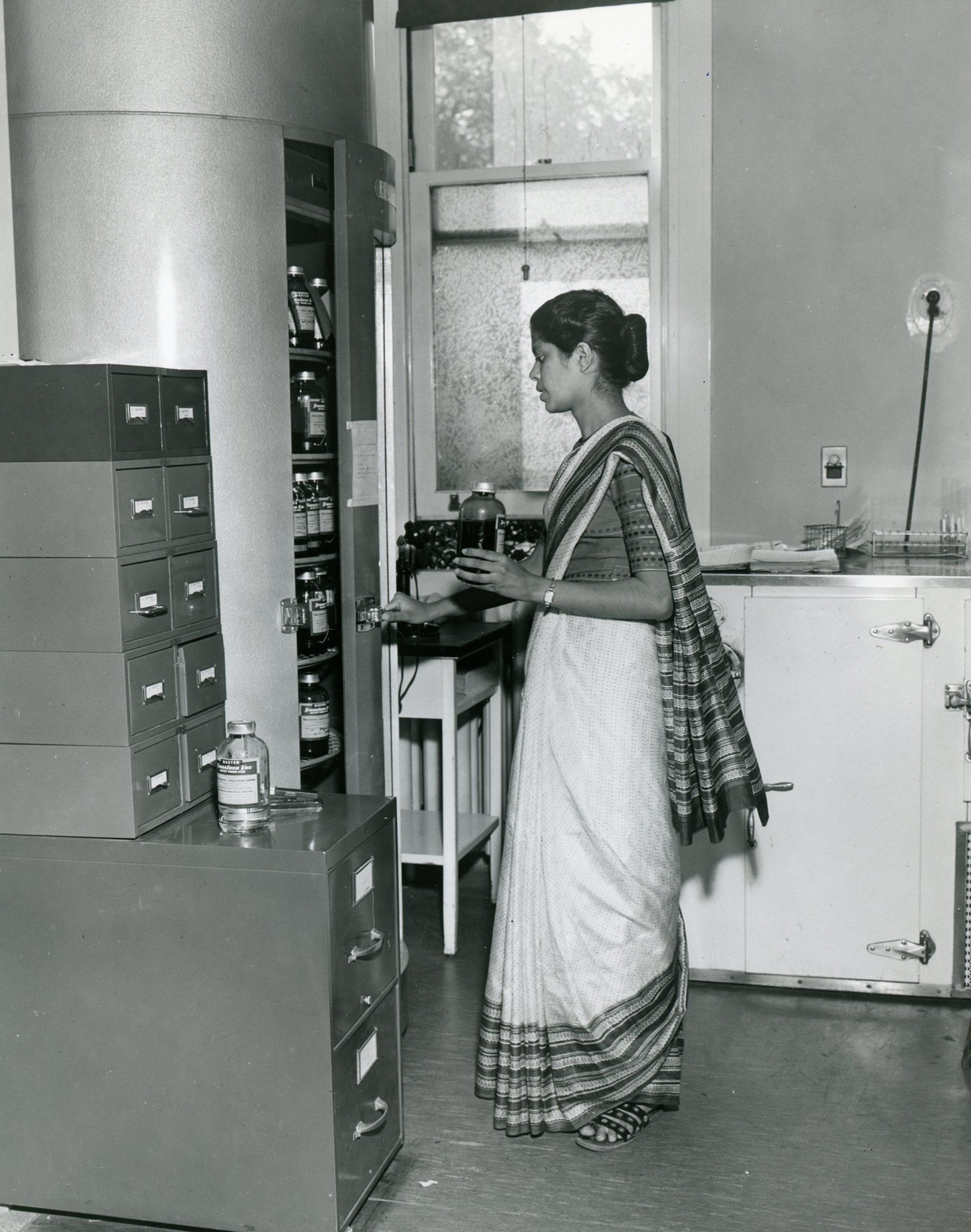 Photo en noir et blanc d’une jeune femme en sari fouillant dans une armoire de matériel médical.