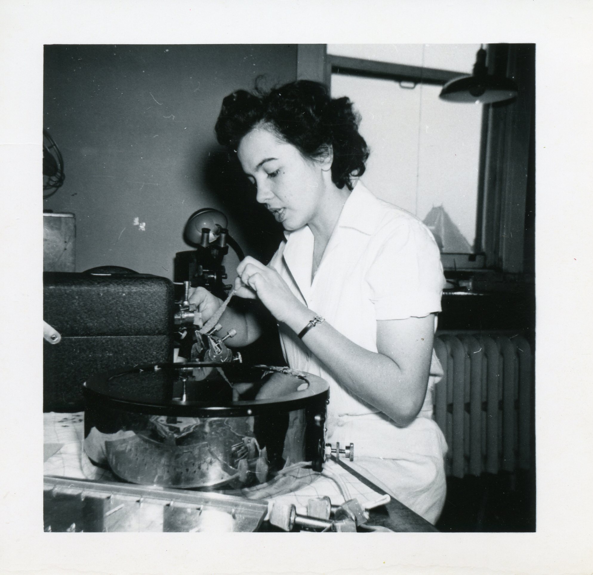 Photo en noir et blanc d’une femme en sarrau de laboratoire vérifie l’état d’équipements scientifiques.