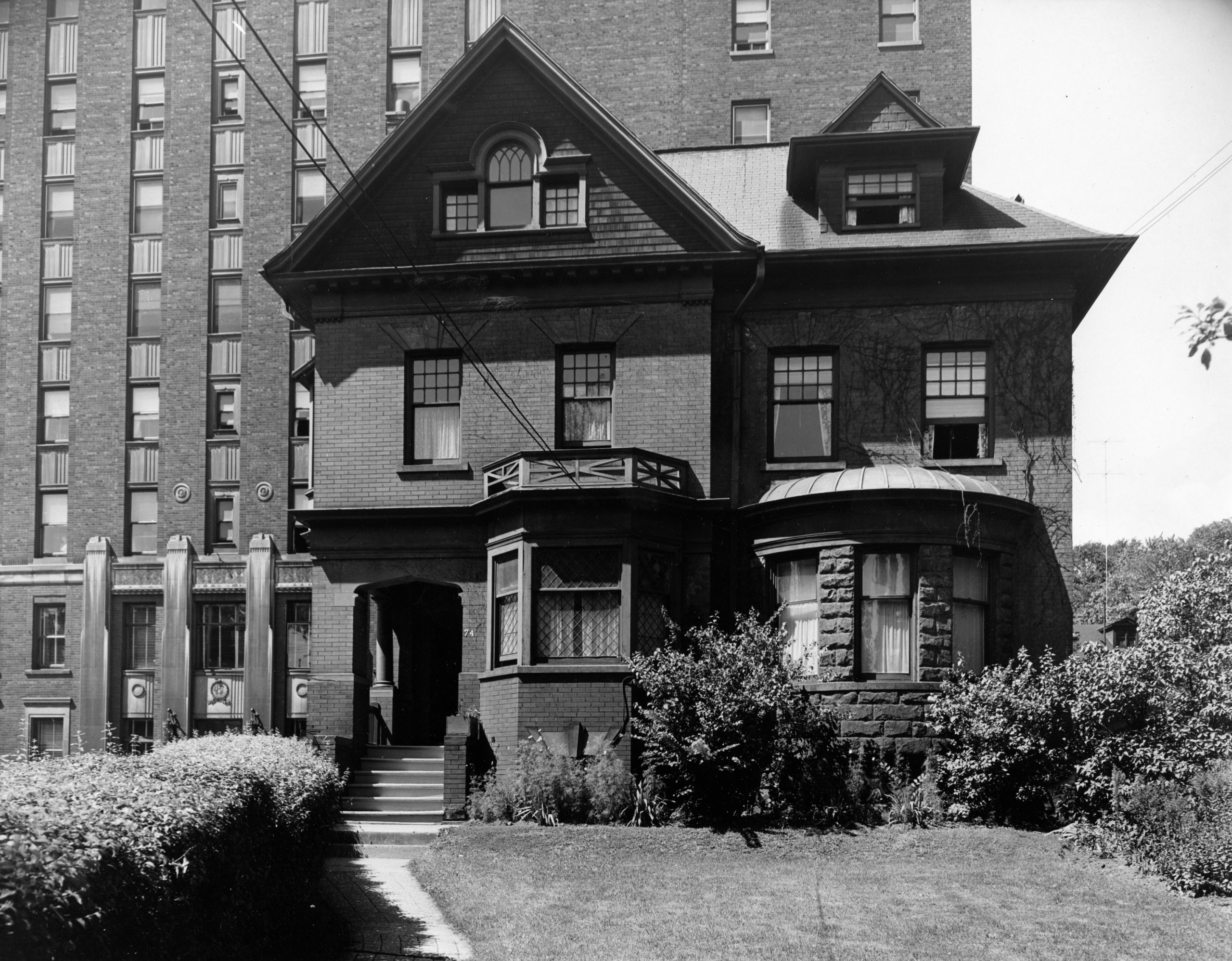 Photo en noir et blanc d’une maison à deux étages. L’hôpital imposant se trouve en toile de fond.