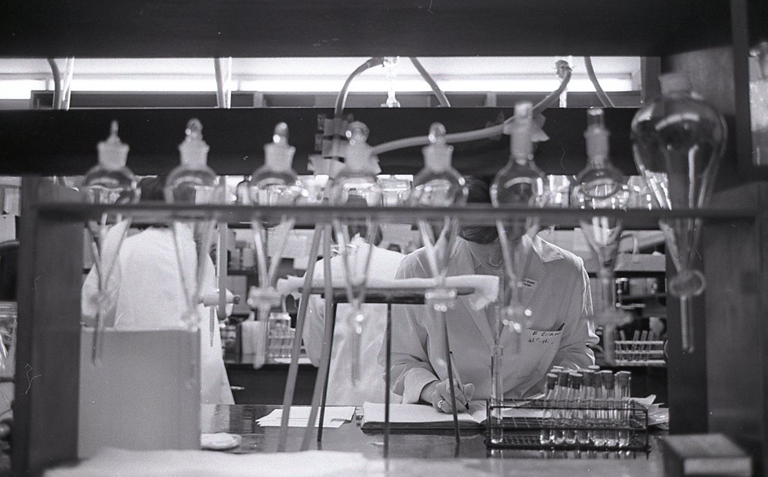 Photo en noir et blanc où l’on voit trois femmes en sarrau de laboratoire derrière une rangée de béchers et d’équipements scientifiques.