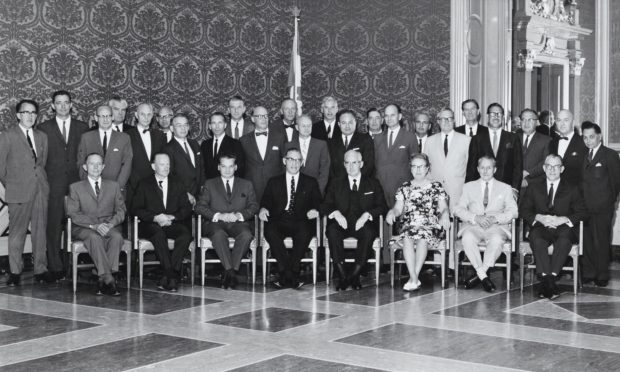 Photo en noir et blanc d’un groupe de personnes élégamment vêtues. Une seule de la trentaine est une femme. Ils posent dans une salle élégante.