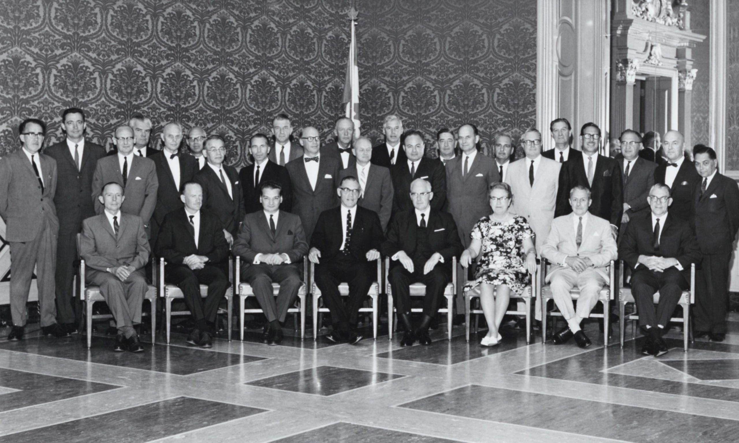 Photo en noir et blanc d’un groupe de personnes élégamment vêtues. Une seule de la trentaine est une femme. Ils posent dans une salle élégante.