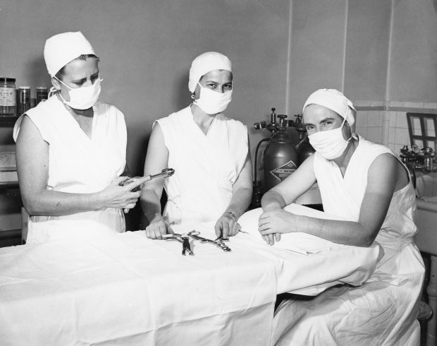 Photo en noir et blanc de trois personnes en blouses chirurgicales posant près d’une table d’opération. Ils ont les yeux souriants.