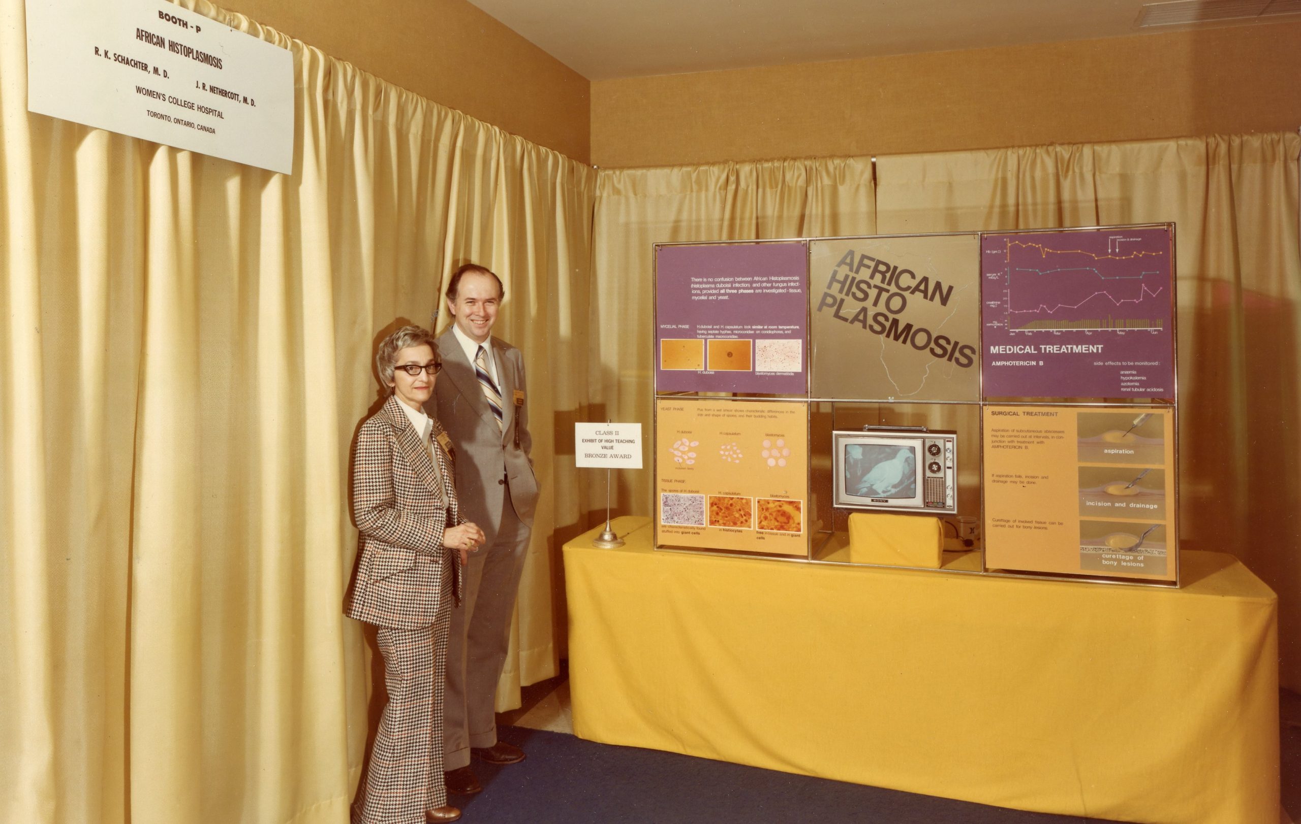 Photo en couleur teintée de jaune d’un homme en complet et d’une femme en tailleur se tenant à côté d’une table d’exposition munie d’un téléviseur et de tableaux d’affichage.