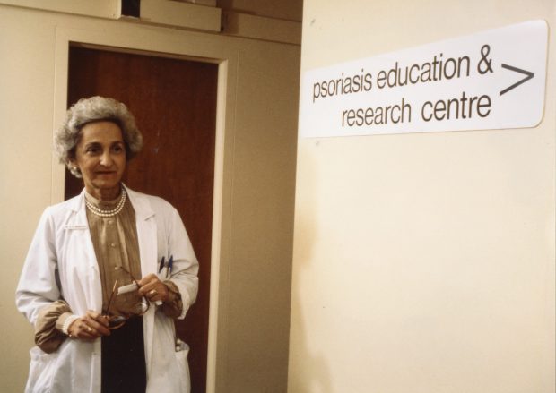 Photo en couleur d’une femme d’âge mûr portant un sarrau de laboratoire et regardant le long d’un corridor près d’une enseigne annonçant le « Psoriasis Education & Research Centre ».