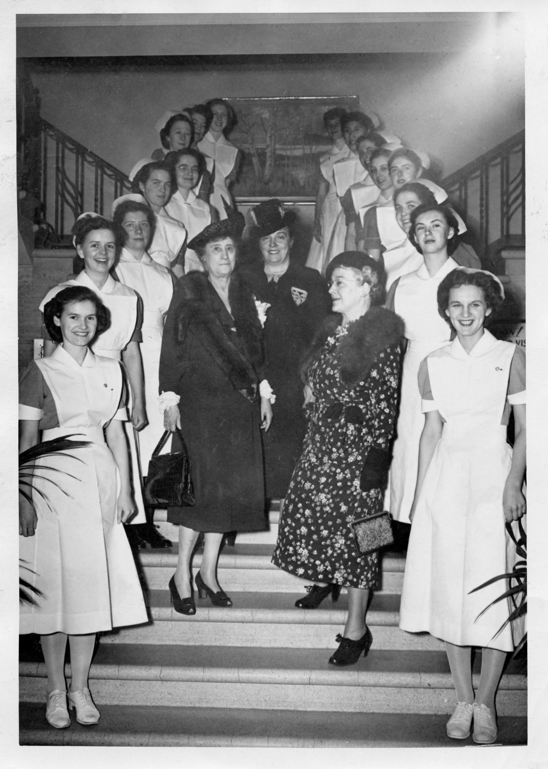 Photo en noir et blanc d’un groupe de femmes sur un escalier. Deux rangées de jeunes femmes en uniforme d’infirmière longent les côtés, alors que trois femmes plus âgées élégamment vêtues se tiennent au centre.