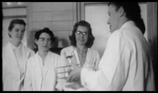 Image d’une vidéo en noir et blanc où des femmes en sarrau de laboratoire se font enseigner par une femme plus âgée.