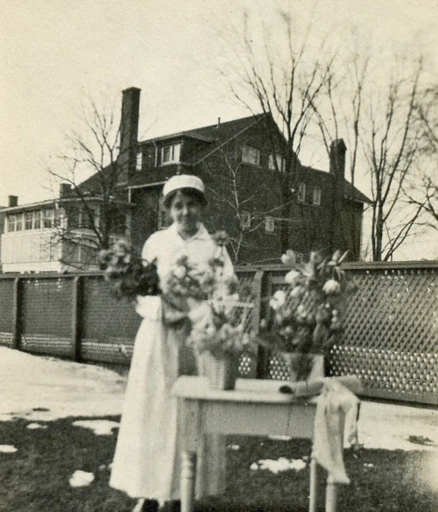 Une jeune femme souriante en uniforme d’infirmière tient un bouquet. Elle est dehors, et près d’elle se trouve une table avec des vases et un rouleau de papier avec ruban.
