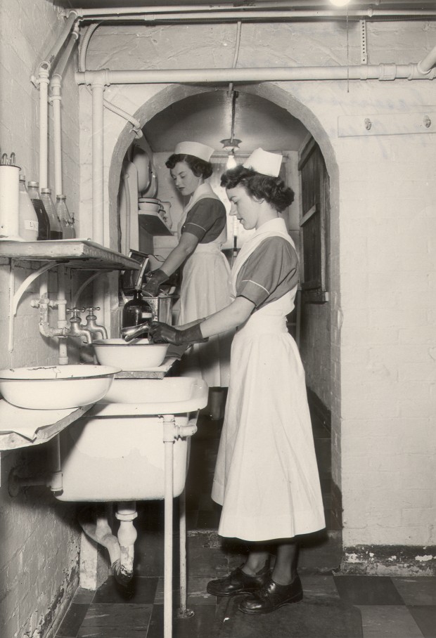 Deux femmes en uniforme d’infirmière portant des gants et faisant du nettoyage devant une série d’éviers.