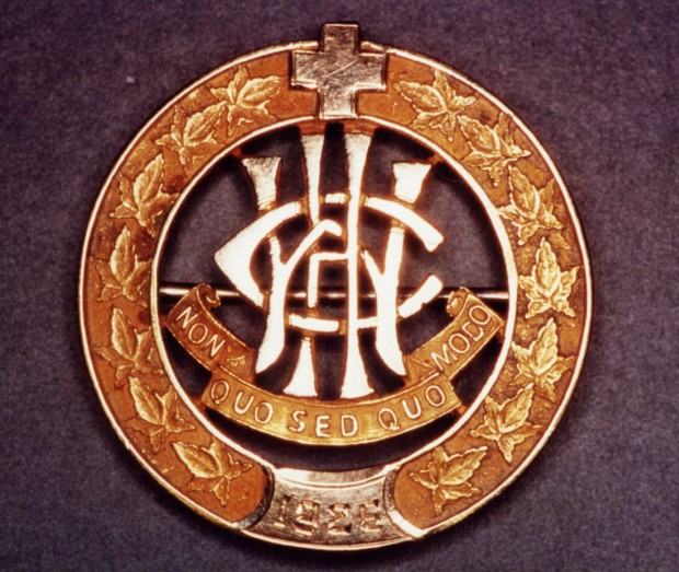Épingle circulaire, bordée de feuilles et couronnée d’une croix. Les lettres W, C et H sont entrelacées au-dessus de la devise.