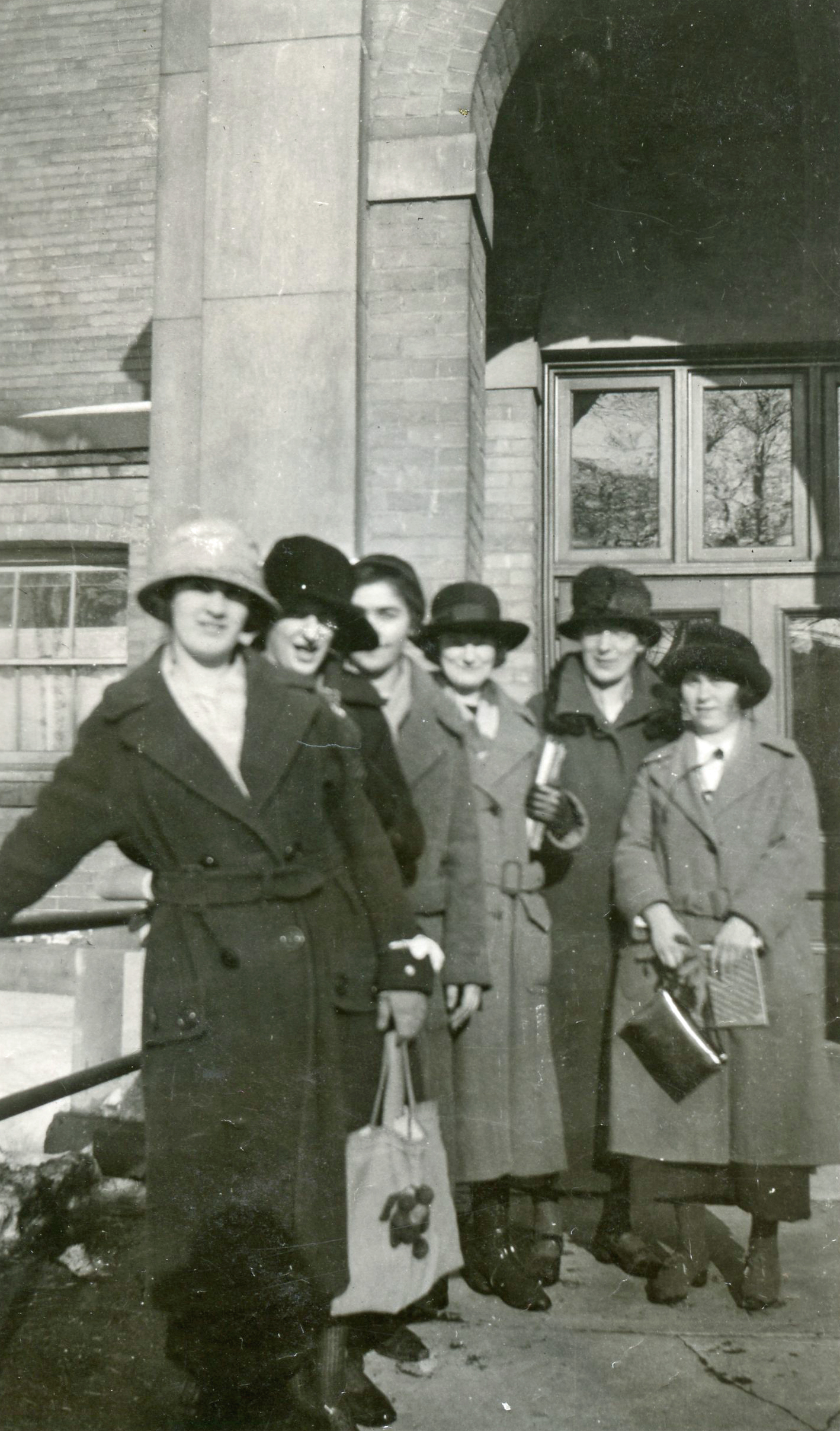 Sept femmes en manteau long et chapeau se tiennent devant une entrée voûtée.