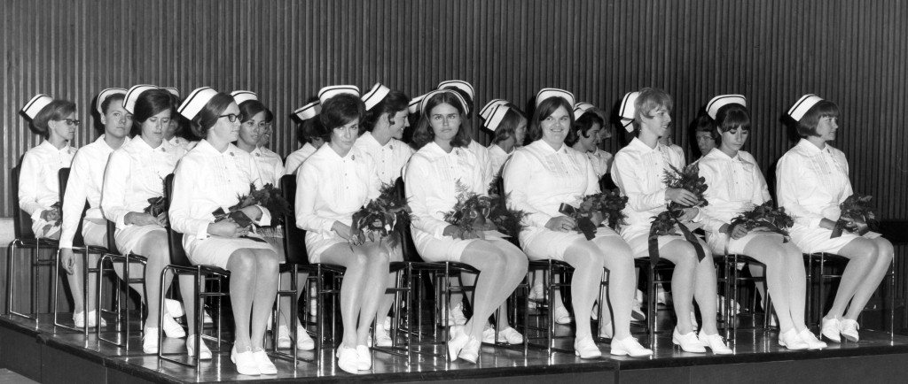 Plus de 20 femmes en uniforme d’infirmière complet assises sur une scène et tenant des fleurs. 