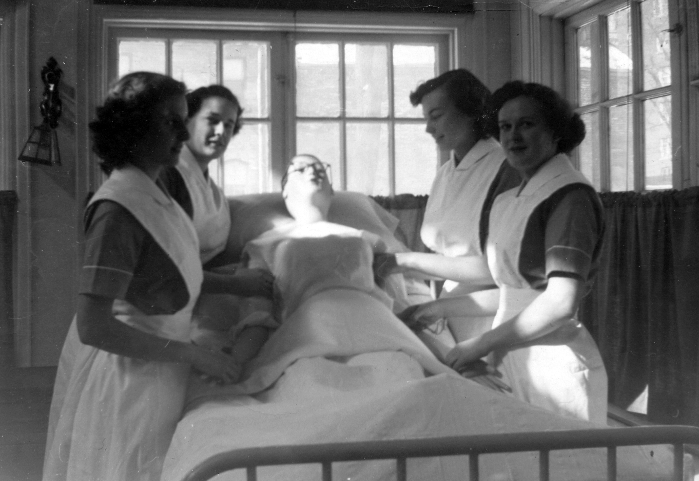 Quatre femmes en uniforme d’infirmière sans coiffe entourent un lit et tiennent un mannequin.