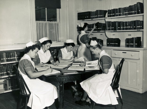 Quatre femmes assises à une table couverte de livres. Une cinquième tend la main vers une étagère.