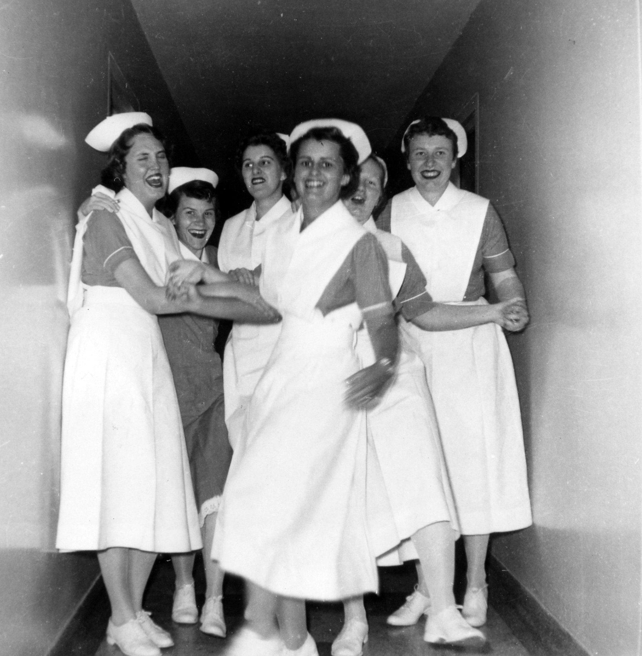 Six femmes en uniforme d’infirmière rient et gesticulent dans un corridor étroit.