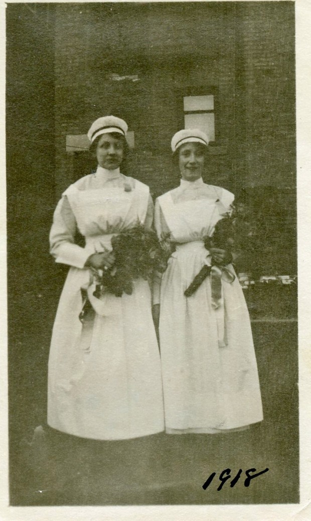 Deux femmes en uniforme d’infirmière se tiennent devant un immeuble de briques, tenant des bouquets et souriant.