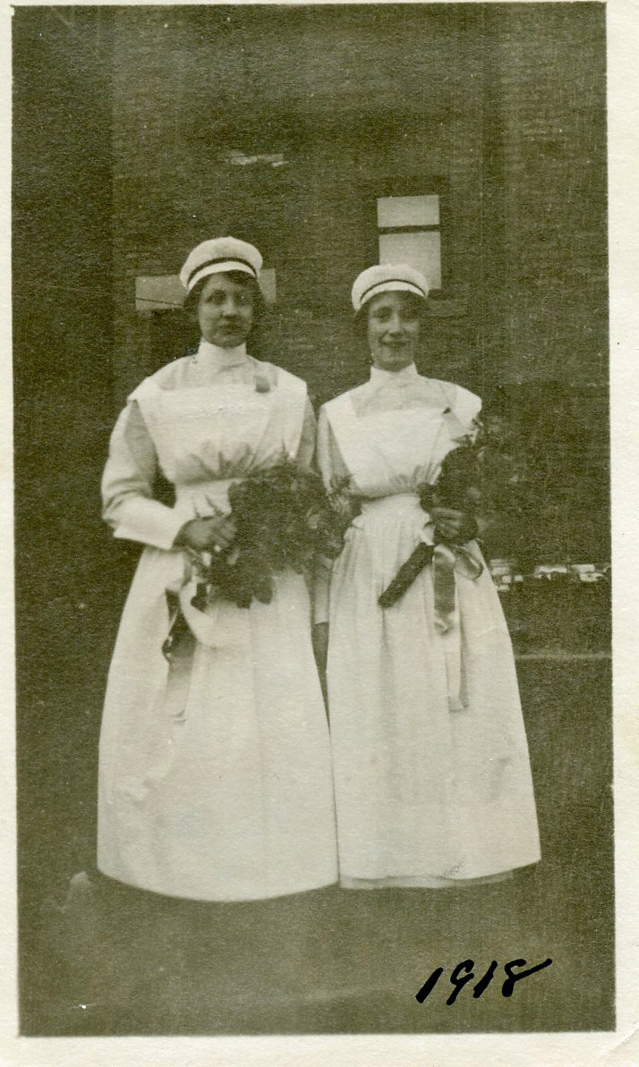Deux femmes en uniforme d’infirmière se tiennent devant un immeuble de briques, tenant des bouquets et souriant.