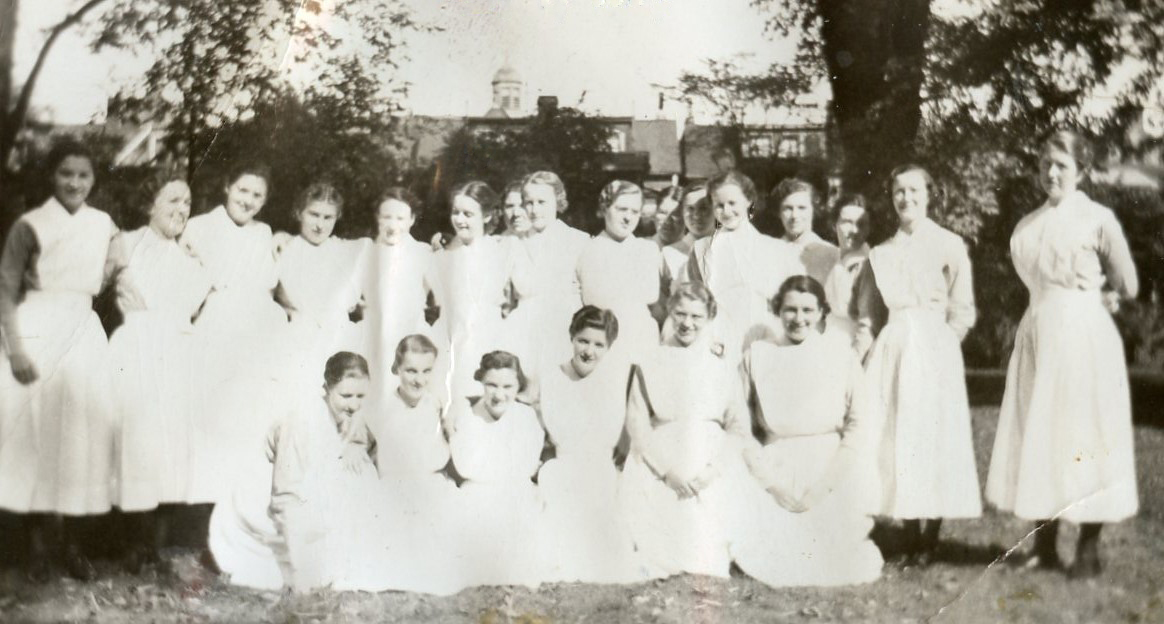 Plus de 20 femmes en uniforme d’infirmière, mais sans la coiffe, sourient à l’extérieur, debout ou à genoux.