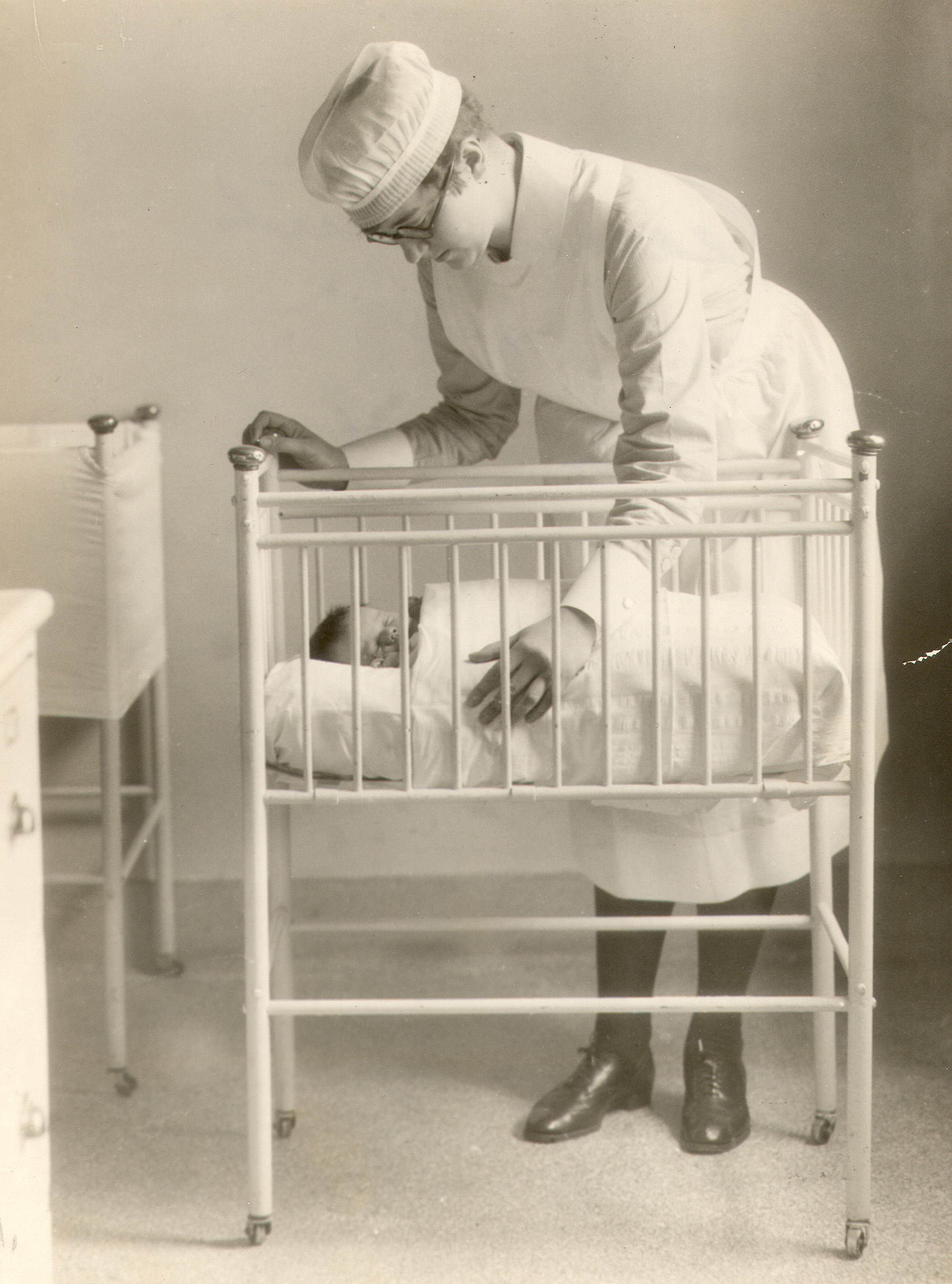 Une jeune femme se penche sur un berceau où se trouve un bébé. Elle porte un uniforme d’infirmière.