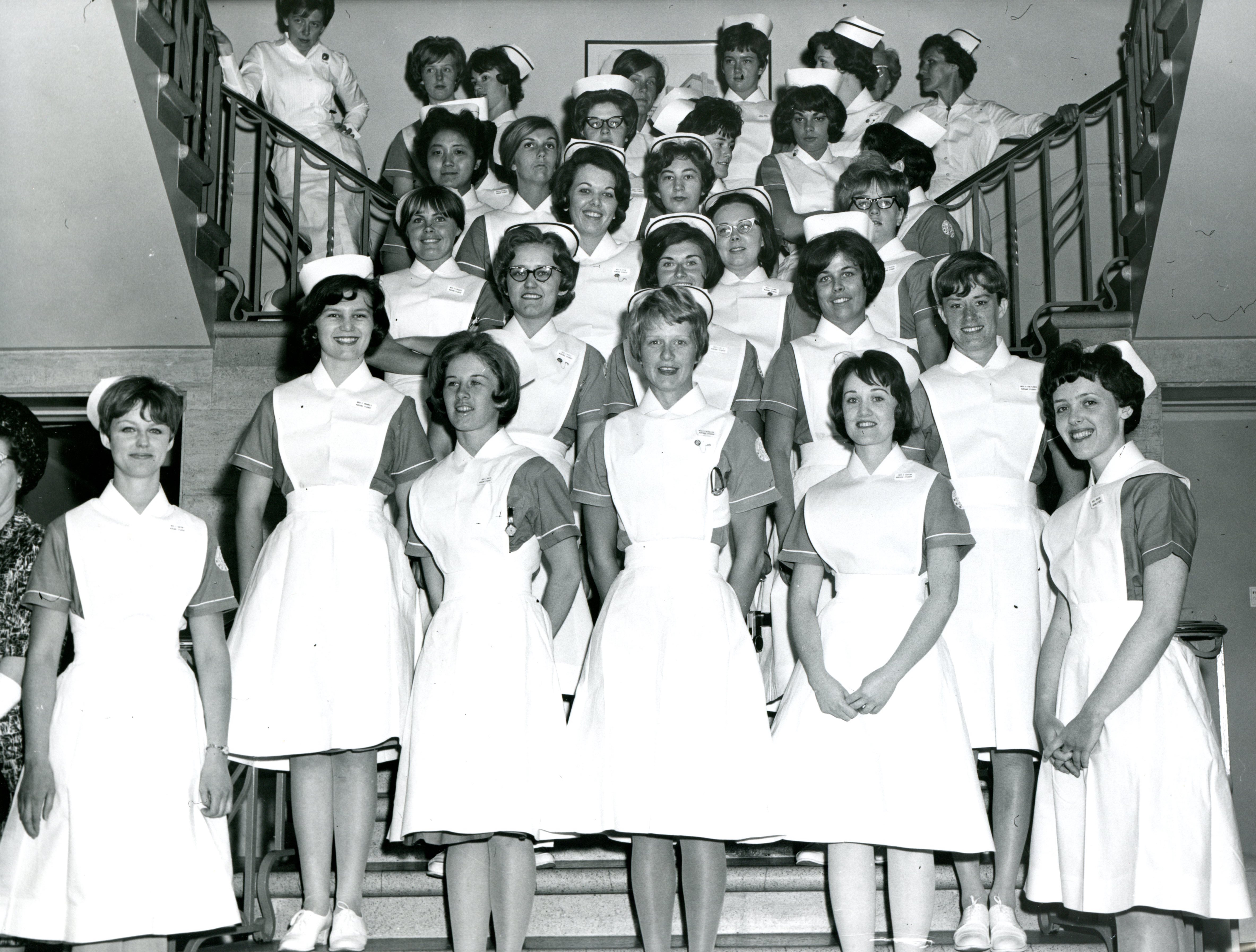 Un grand groupe de femmes pose pour un photographe sur un escalier. Elles portent des uniformes d’infirmière avec coiffes.