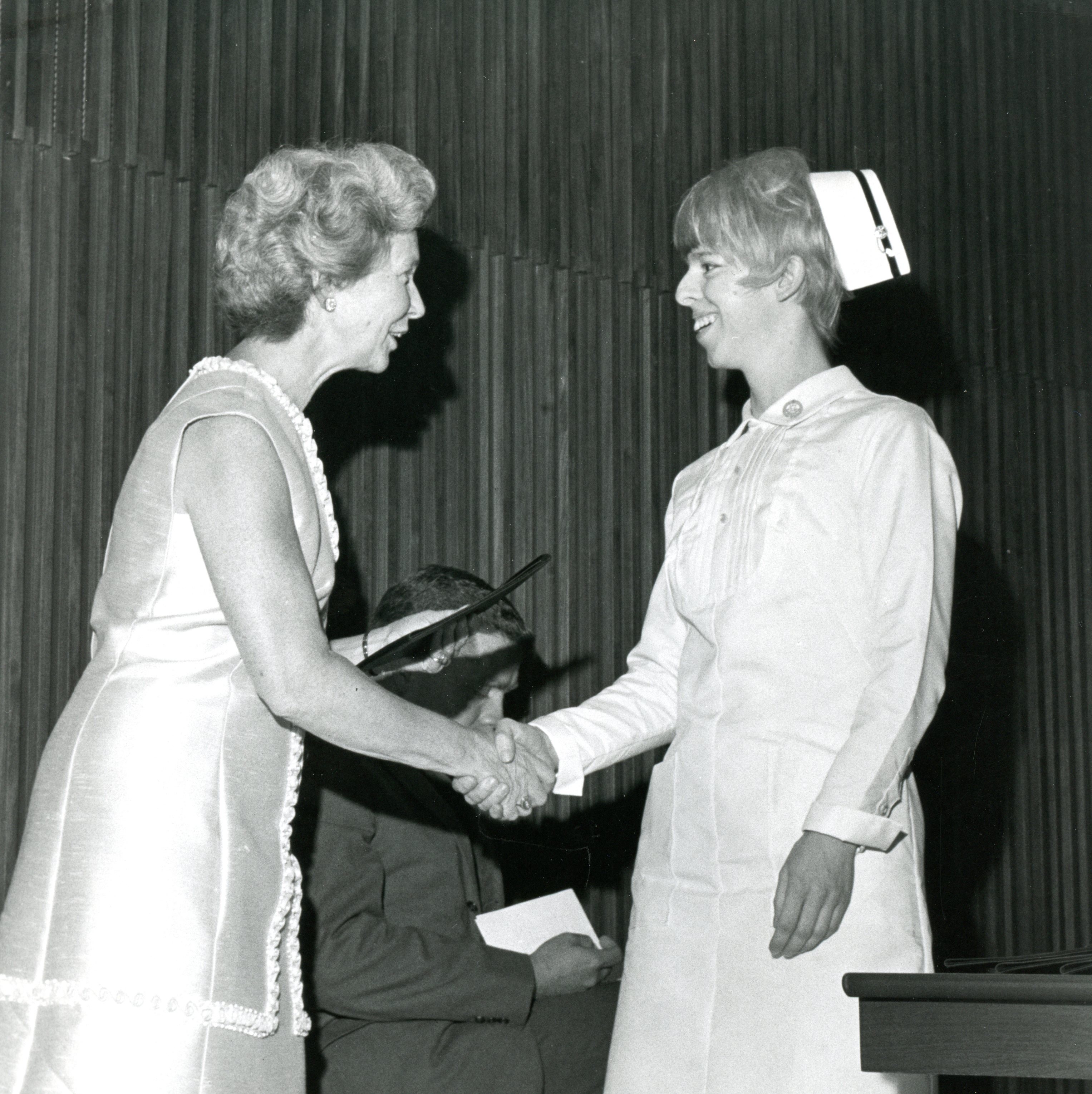 Deux femmes souriantes se serrent la main. L’une d’entre elles porte une robe habillée et l’autre un uniforme d’infirmière avec coiffe. Un homme se tient à l’arrière-plan.