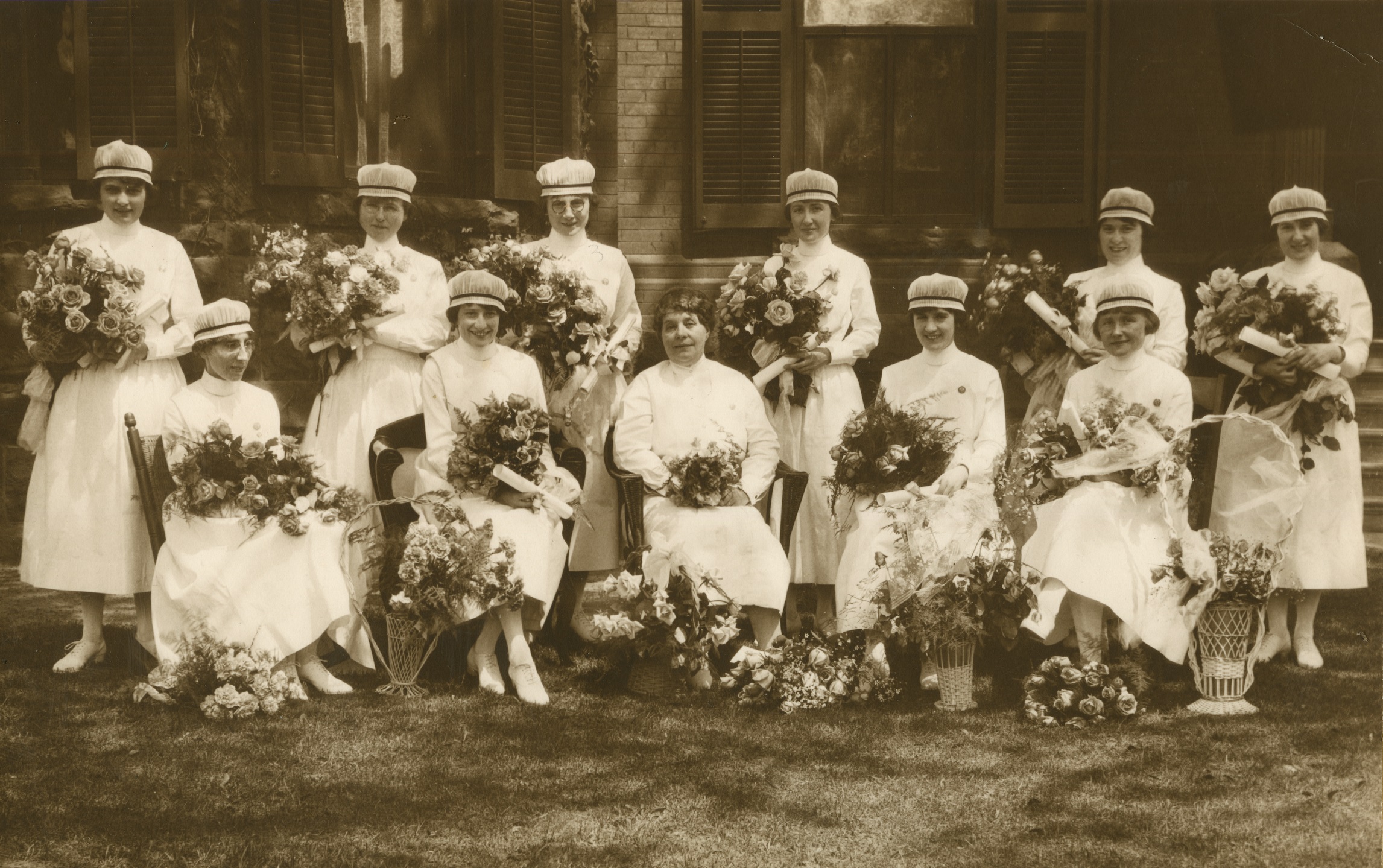 Onze femmes souriantes posent officiellement avec des bouquets de fleurs. Toutes les femmes sauf une portent une coiffe d’infirmière.