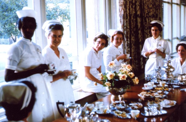 Six jeunes femmes vêtues d’uniformes d’infirmière se tiennent devant une table décorée remplie de tasses à thé et de biscuits dans une pièce avec de grandes fenêtres.