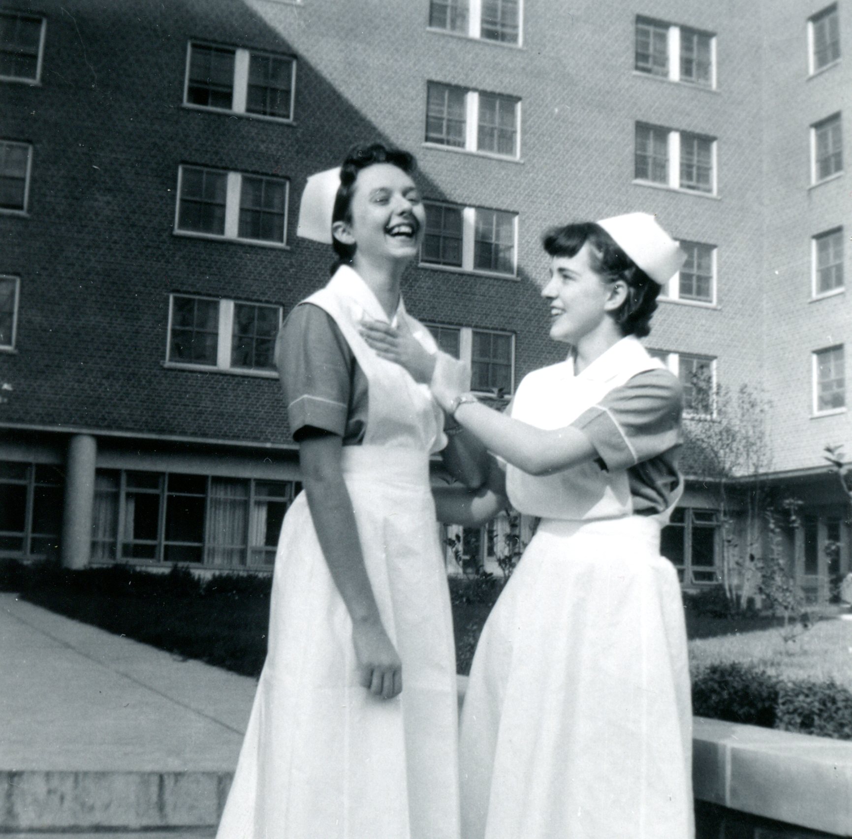 Deux jeunes femmes portant l’uniforme et la coiffe d’infirmière plaisantent affectueusement devant un grand immeuble en briques.