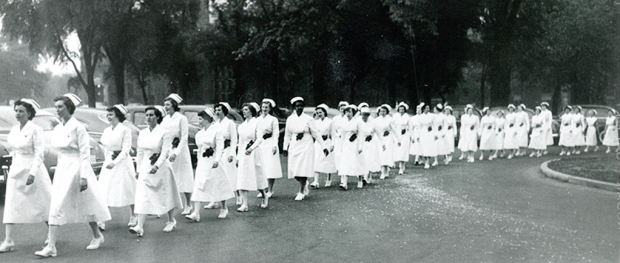 Plus de 30 femmes marchent en rangée, portant uniforme, coiffe et souliers blancs.