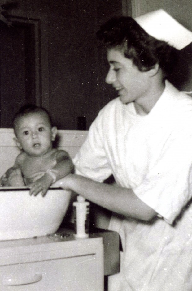 Une jeune femme souriante, portant uniforme et coiffe d’infirmière, donne un bain à un bébé dans une cuvette.