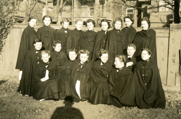 Dix-huit jeunes femmes posent pour une photo. Elles portent des capes d’infirmière fermées et sont dehors devant une clotûre. On peut voir l’ombre du photographe au sol.