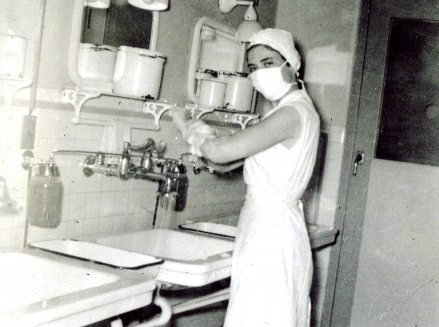 Une jeune femme se savonne jusqu’aux coudes au-dessus d’un évier. Elle regarde l’appareil photo, et l’on voit qu’elle sourit malgré le masque chirurgical. Elle porte divers éléments de l’uniforme d’infirmière, bien qu’elle n’ait pas de manches. Ses cheveux sont complètement couverts par un bonnet protecteur.