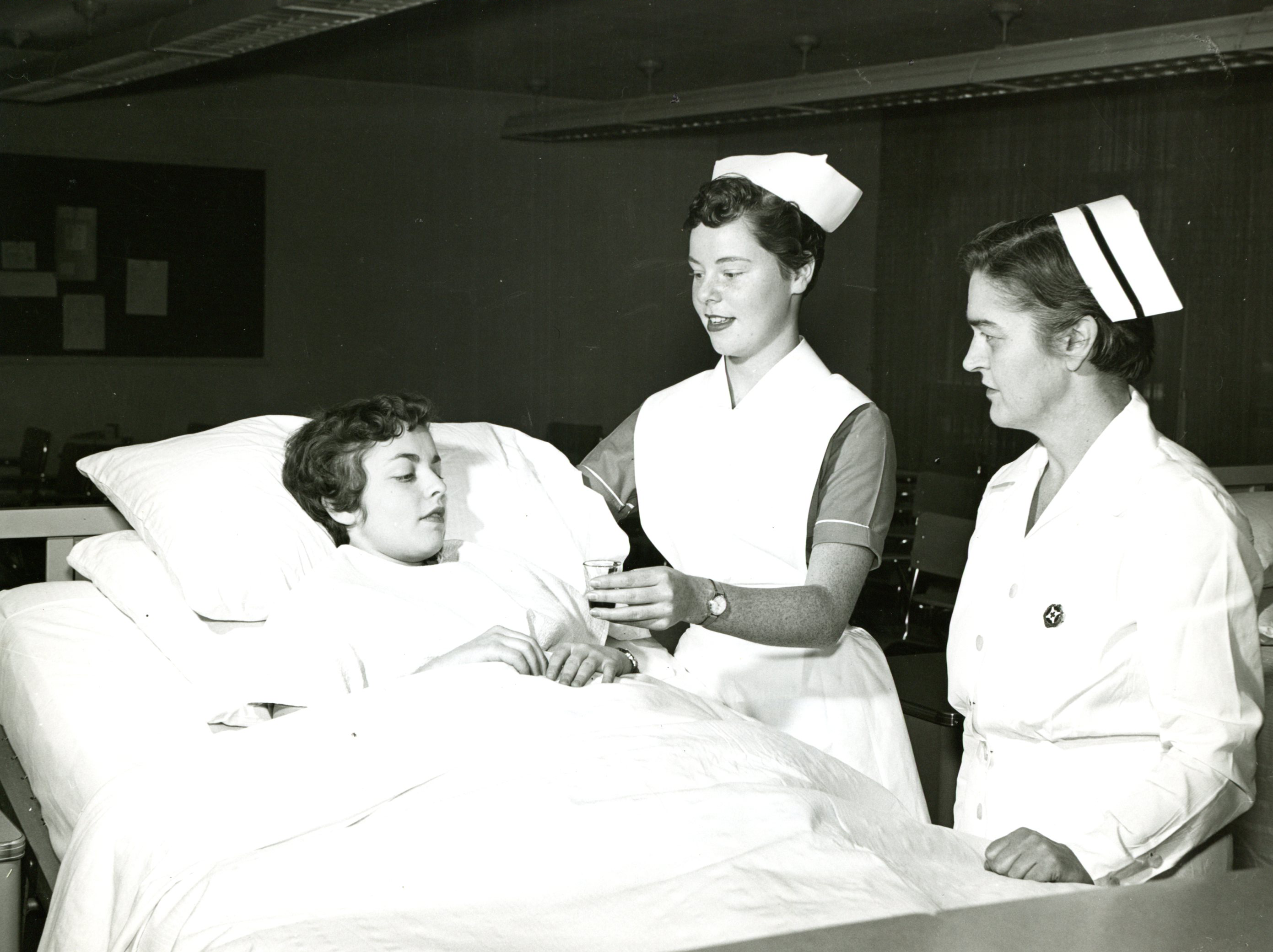Une jeune femme en chemise d’hôpital se repose au lit pendant qu’une autre, vêtue d’un uniforme d’infirmière, lui tend un petit verre. Une autre femme portant un uniforme et une coiffe d’infirmière observe.