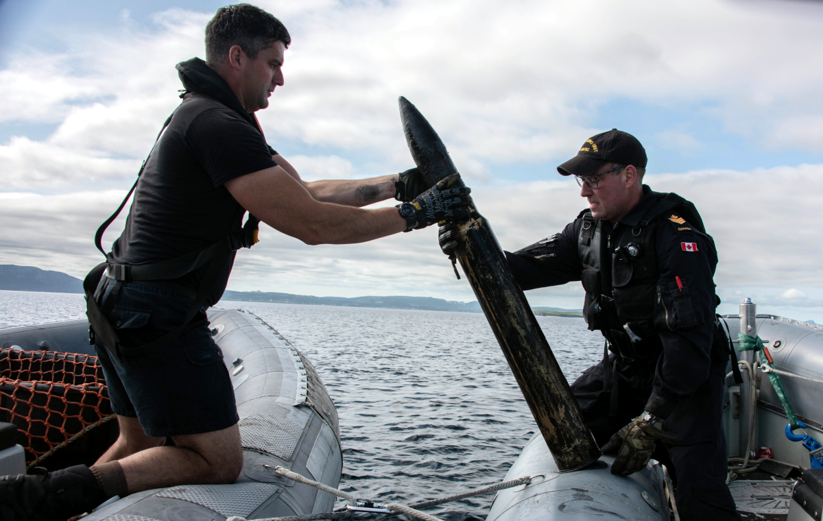 Deux plongeurs de la marine soulèvent un vieil obus d’artillerie entre deux bateaux