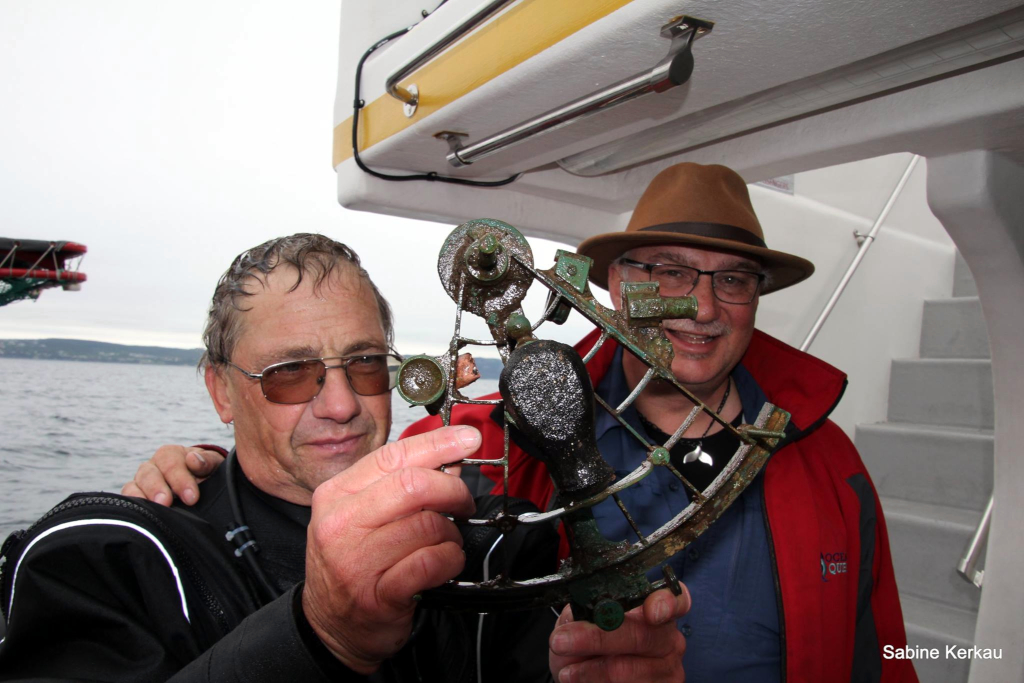 Un plongeur tient un sextant en laiton mouillé pendant qu’un autre homme regarde en souriant