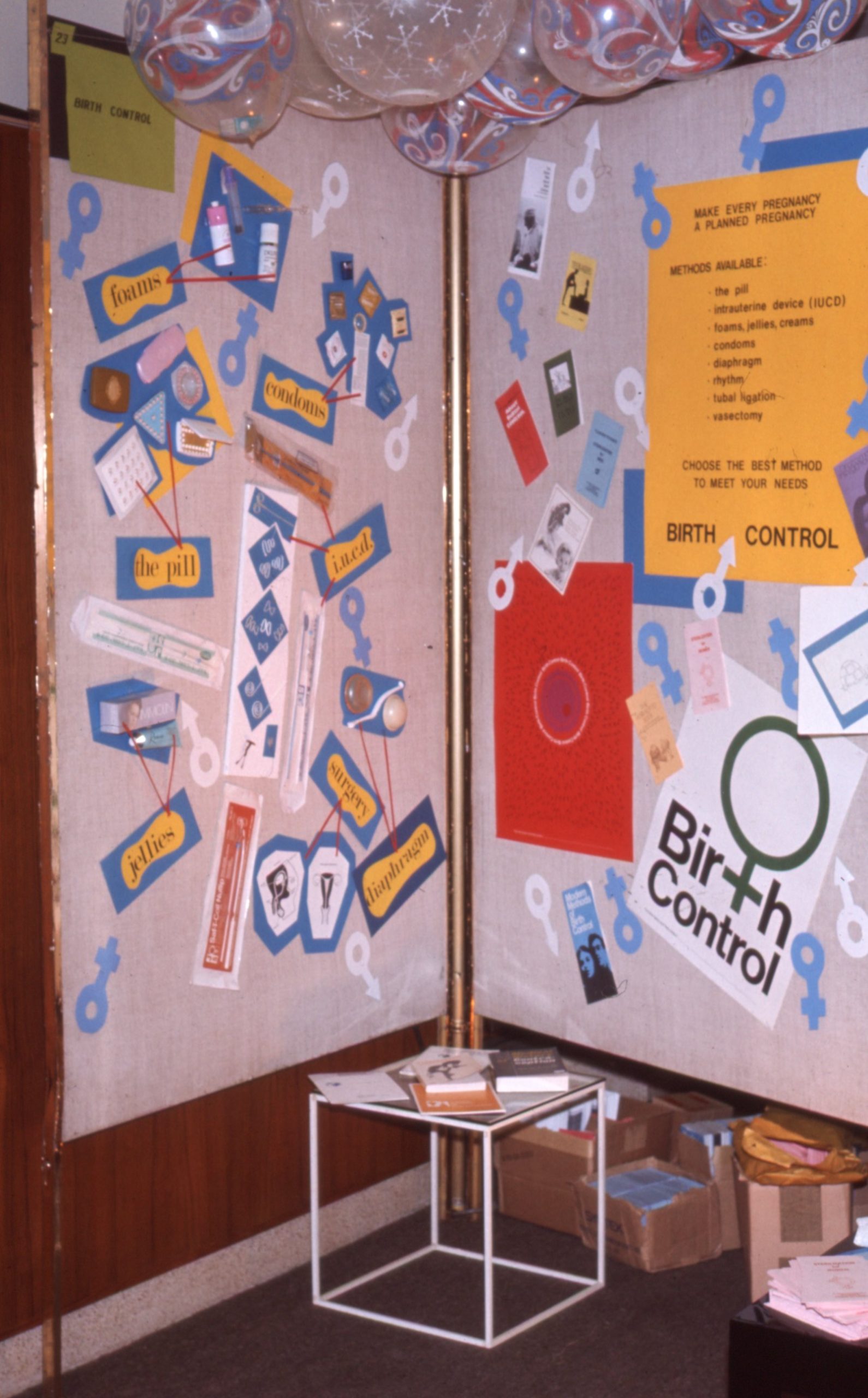 Présentoir de produits contraceptifs consistant en deux panneaux verticaux formant un coin. Les panneaux sont couverts d’une variété de découpages de papier colorés, d’affiches et de produits contraceptifs.